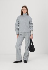 Grå fleece turtleneck genser, vide grå bukser med sømdetaljer, og en svart vevd håndveske. Modellen står mot en lys bakgrunn.