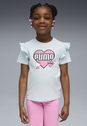 Junges Mädchen mit geflochtenen Zöpfen, das ein weißes Puma Hello Kitty T-Shirt und rosa Hosen vor einem grauen Hintergrund trägt.