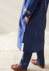 Person iført en blå broderet lang frakke med hænderne i lommerne, blå bukser og brune mokkasiner med kvaster, der står på et lyst tæppe.