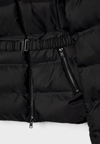 Czarna pikowana kurtka z regulowanym pasem, podwójnymi kieszeniami zamykanymi na zamek i gładką matową teksturą. Dodatki obejmują płaski detal z klamrą.