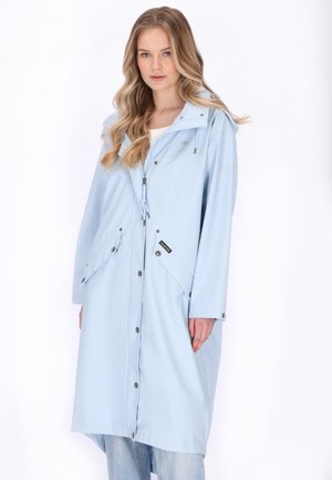 Hosszú, hullámos szőke hajú nő világoskék kapucnis esőkabátot visel, melyen patentos zsebek és cipzár találhatók, fehér háttér előtt áll.