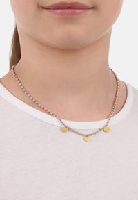 Bunte Perlenhalskette mit goldenen Herzanhängern, die rosa, türkis und weiße Akzente aufweist, auf unbedeckter Haut über einem weißen Shirt getragen.
