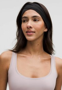 Un bandeau en tissu noir avec un design torsadé, porté sur des cheveux ondulés et foncés. Le sujet porte un haut sans manches rose clair.