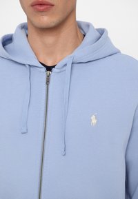 Ljusblå zip-up hoodie i mjukt tyg, med en framficka, dragsnoddar och en liten broderad logotyp i en kontrasterande färg.