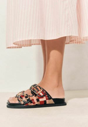 Füße in offenen Sandalen mit roten, schwarzen und beigen gewebten Riemen und Schnallen, unter einem hellrosa gestreiften Rock.