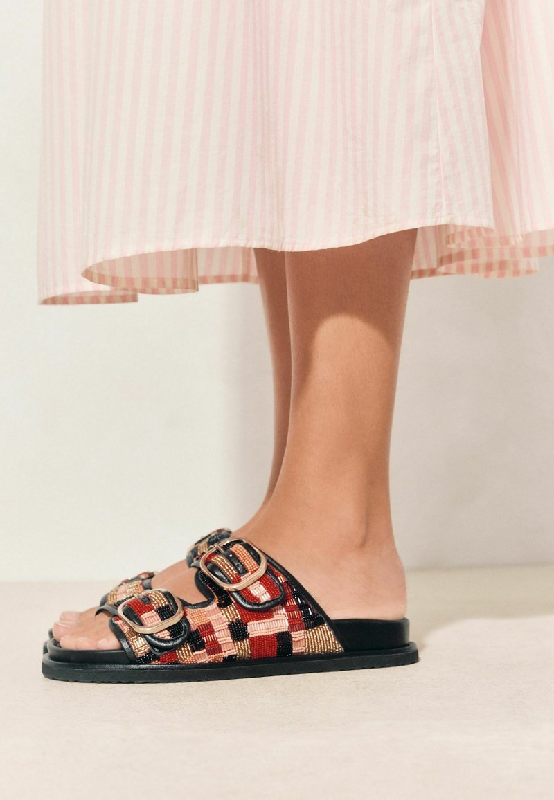 Piedi che indossano sandali con punta aperta con cinturini intrecciati rossi, neri e beige e fibbie, sotto una gonna a righe rosa chiaro.