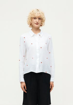 Persoon met krullend blond haar, een witte blouse met knopen met kleine rode hartjespatronen en een zwarte broek met wijde pijpen.