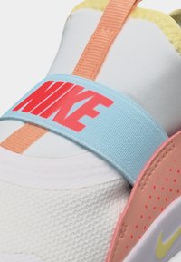 Κοντινό πλάνο λευκού αθλητικού παπουτσιού με πορτοκαλί λουράκι, μπλε ταινία με τη λέξη "NIKE" σε κόκκινο και ροζ-κίτρινες λεπτομέρειες στη σόλα.