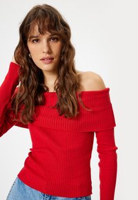 Top rosso a coste con scollo off-the-shoulder e maniche lunghe. Il tessuto morbido, il design aderente e la finitura testurizzata creano una silhouette che valorizza la figura.