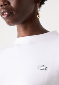 White cotton shirt with a round neck, featuring a small black and white embroidered crocodile logo on the chest. Textured surface.
