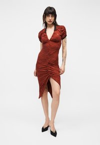 Γυναίκα που φοράει φόρεμα σε απόχρωση σκουριάς με ρυτιδώσεις, γιακά και μαύρο περιθώριο, στέκεται με ένα πόδι ελαφρώς μπροστά και τα χέρια χαλαρωμένα στο πλάι.