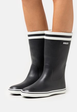 Botas de lluvia de goma negras con ribete y franjas blancas en la parte superior, con una textura suave y punta redondeada. Etiqueta de la marca en el lado.