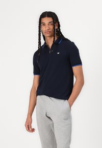 Polo-shirt bleu marine à manches courtes, col zippé et accents bleus contrastants. Associé à un pantalon de jogging gris clair.