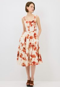 Vestido con estampado floral en color crema con acentos rojos. Presenta un escote cuadrado, diseño sin mangas, bolsillos y una falda acampanada. Lleva puestos unos tacones marrones.