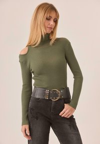 Suéter de canalé verde oliva con cuello simulado y un escote en un hombro, combinado con un cinturón de cuero negro que presenta herrajes dorados y tachuelas.