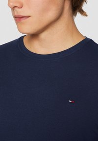 Námorně modré bavlněné tričko s kruhovým výstřihem, které má malý červený, bílý a modrý logotyp na hrudi. Hladký povrch.