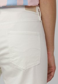 Les jeans en denim blanc présentent une poche arrière unique avec un détail de couture courbe. Le tissu est lisse avec une finition propre.