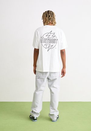 White cotton t-shirt featuring a black "DETROIT" graphic and lightning bolt design on the back, paired with light grey trousers.