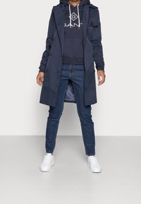 Cappotto trench navy con design doppiopetto, felpa abbinata, jeans di denim e sneakers bianche. Presenta dettagli a bottoni e spacchi laterali.