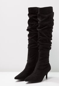 Botas altas de ante negro con diseño fruncido, punta afilada y tacones de aguja. Cuenta con una cremallera lateral para facilitar su uso.