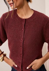 Bordový pletený kardigan s okrúhlym výstrihom, krátkymi rukávmi a zapínaním na gombíky vpredu. Má jemnú textúru a voľnejší strih.