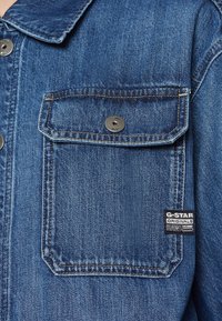 Veste en denim avec une poche avant, délavage bleu clair, fermeture à boutons, couture contrastante et étiquette G-Star RAW sur le côté gauche.