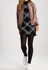 Brązowa satynowa kurtka bomberka na czarno-białej sukience w wzory, zestawiona z czarnymi rajstopami i białymi trampkami.