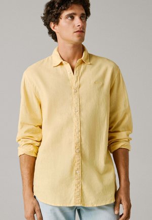 Młody mężczyzna w jasnożółtej koszuli na guziki z długim rękawem, z podwiniętymi rękawami, stojący na tle jednolitego jasnego tła.