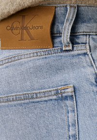 Jeans din denim albastru deschis, cu o etichetă din piele maro pe care este embosat "Calvin Klein Jeans". Cusături de contrast și un design standard al buzunarelor sunt vizibile.