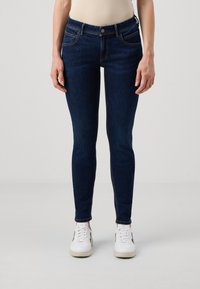 Mörkblå slim fit jeans med medelhög midja, med fem fickor och kontrasterande sömmar. Matchas med vita sneakers för kontrast.