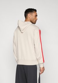 Felpa con cappuccio beige con strisce rosse laterali, caratterizzata da una vestibilità rilassata e polsini a coste. Tessuto morbido con una texture liscia.