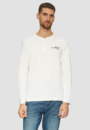 Langärmliges weißes Henley-Shirt aus weichem Stoff, mit einem Knopfkragen und gedrucktem Text auf der linken Brustseite. Kombiniert mit blauen Jeans.