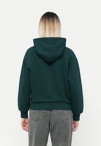 Sweat à capuche vert foncé au fit décontracté, avec des poignets et une taille côtelés. Il possède une grande capuche et un tissu doux et texturé.