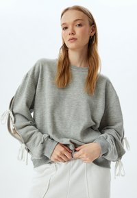 Felpa grigia con maniche lunghe, caratterizzata da dettagli con cordino ai polsini. Tessuto morbido con vestibilità rilassata, abbinata a pantaloni bianchi.