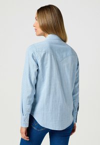 Jasnoniebieska koszula w kratę z długimi rękawami, z guzikami na mankietach, spiczastym kołnierzykiem i detalem w kształcie yoke na plecach, w połączeniu z ciemnoniebieskimi dżinsami.