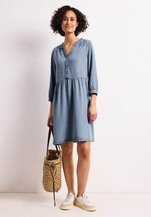 Kobieta z kręconymi włosami, ubrana w niebieską dżinsową sukienkę do kolan, białe trampki, trzymająca plecioną słomianą torbę z paskami ze skóry.