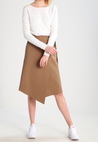 Brown wrap skirt made of smooth fabric, featuring an asymmetrical hemline. Paired with a textured white long-sleeve top and white sneakers.