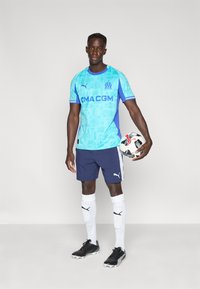 Ανοιχτό μπλε ποδοσφαιρική φανέλα με μπλε λεπτομέρειες και λογότυπο, συνδυασμένη με ναυτικό σορτς και λευκές κάλτσες, κρατώντας μια ασπρόμαυρη μπάλα ποδοσφαίρου.