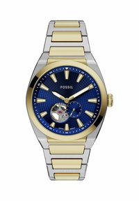 Montre-bracelet Fossil en métal bicolore argent et or avec cadran texturé bleu, index dorés et mouvement mécanique visible à 7 heures.