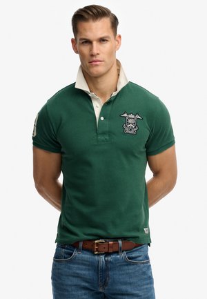 Hombre con camisa polo verde con escudo bordado y cuello beige, vaqueros azules y cinturón marrón, de pie sobre un fondo blanco.