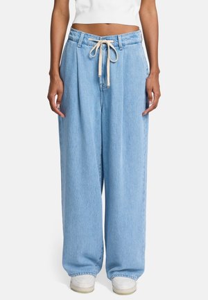 Mujer con vaqueros anchos de pierna suelta azul claro con cordón beige, top blanco corto y zapatillas blancas, de pie frente a un fondo liso.