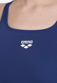 Marineblauer Badeanzug mit einem Rundhalsausschnitt und dünnen Trägern. Verfügt über ein weißes "arena" Logo und ein Diamantmuster unterhalb des Logos.