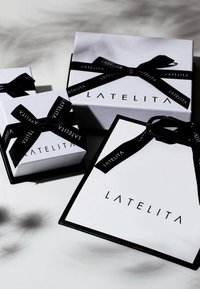 Három fehér ajándékdoboz fekete szalaggal, amelyek a "LATELITA" logót viselik. A legnagyobb doboz négyzet alakú, míg a kisebbek téglalap formájúak.