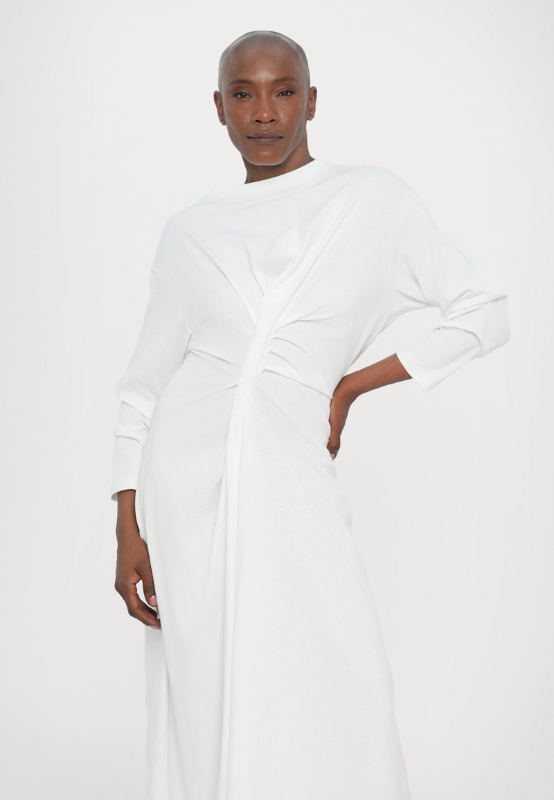 Witte lange mouwen jurk van zachte stof, met een gedraaid ontwerp bij de taille en een vloeiende silhouet. Gladde textuur en minimale details.