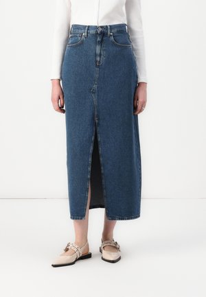 Kvinde iført en højtdækkende blå denim midi-nederdel med en frontslids, kombineret med beige spidse flade sko og en hvid ribbet langærmet top.
