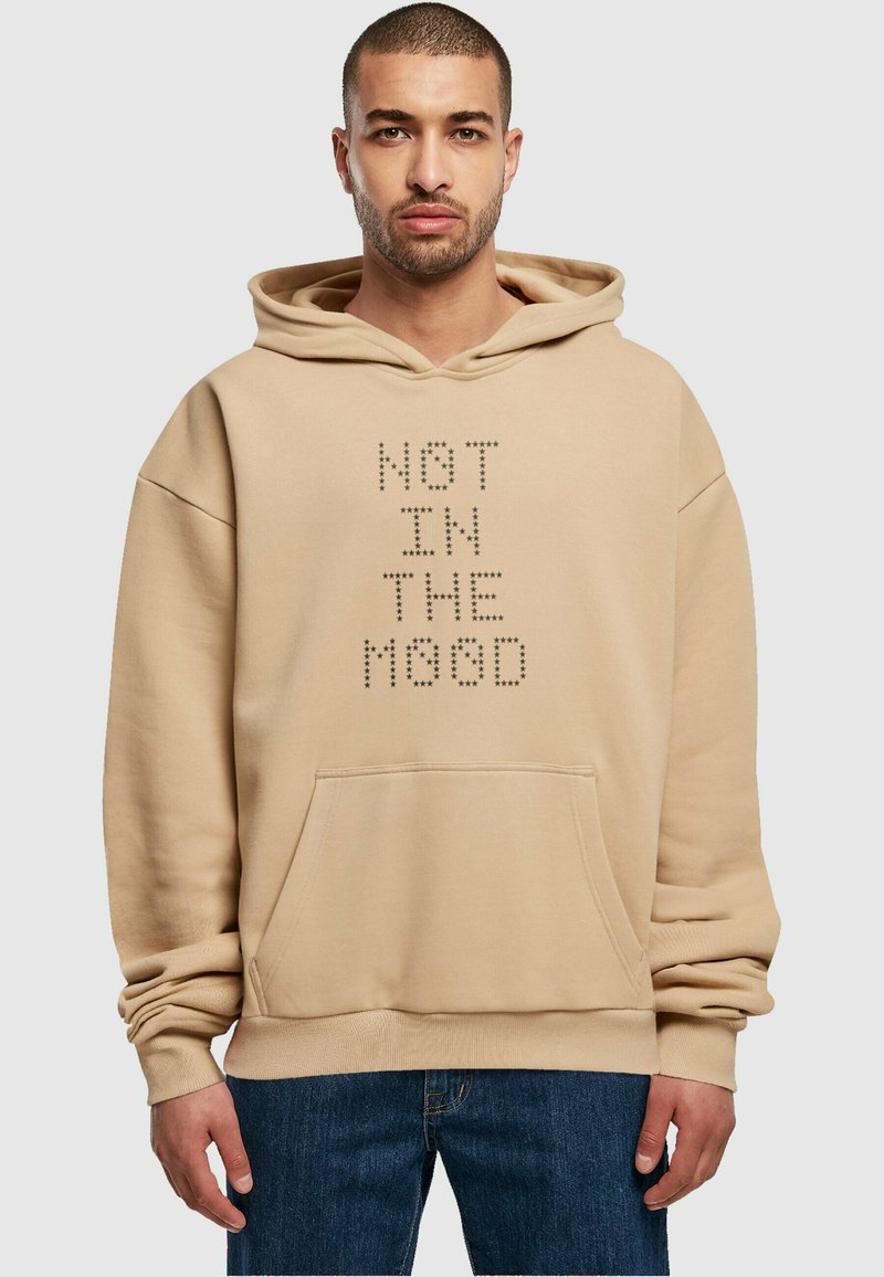 Felpa beige con tasca a marsupio, caratterizzata dal testo "NOT IN THE MOOD" in strass neri. Il tessuto appare morbido con una vestibilità rilassata.