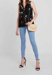 Žena ve černé květované tílko bez rukávů, světle modrých skinny jeans, černých sandálech na vysokém podpatku a držící béžovou kabelku přes rameno.