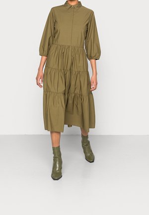 Frau trägt ein olivgrünes, gestuftes Midikleid mit dreiviertellangen Puffärmeln und passenden, strukturierten Stiefeletten.