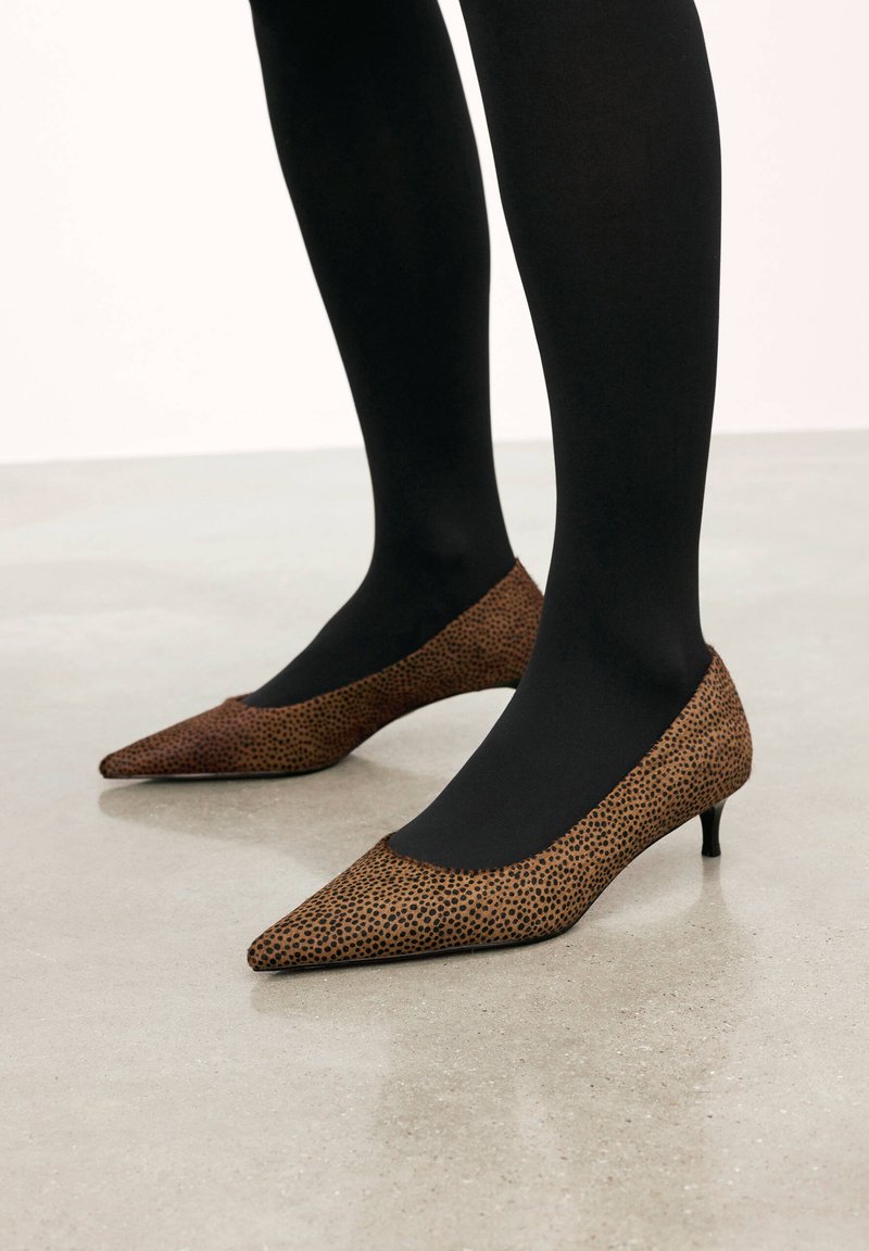 Talons aiguilles à bout pointu en matériau texturé marron avec un motif de pois noirs, avec un petit talon et associées à des collants opaques noirs sur une surface claire.