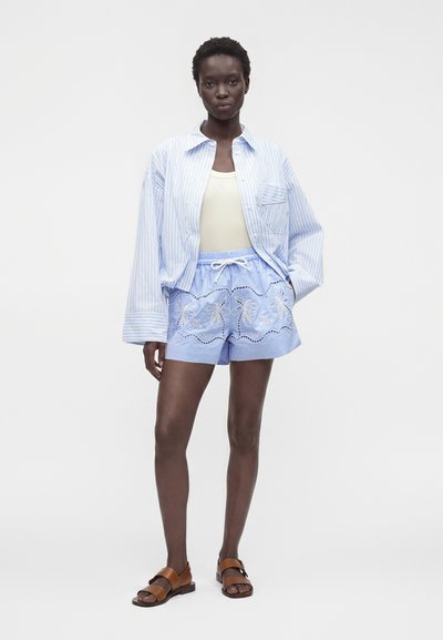 Femme portant une chemise à rayures bleu clair, un débardeur blanc, un short bleu clair brodé et des sandales marron, debout devant un fond uni.